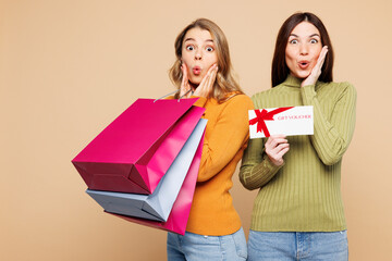 Young amazed friends two women in orange green shirt casual clothes hold shopping package bags gift coupon voucher card for store isolated on plain beige background Black Friday sale buy day concept