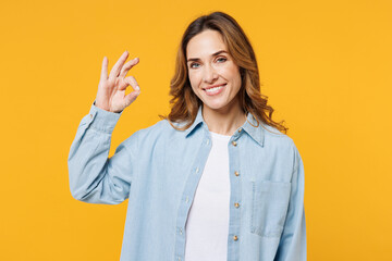 Young smiling happy cheerful woman she wear blue shirt white t-shirt casual clothes showing okay ok gesture looking camera isolated on plain yellow wall background studio portrait. Lifestyle concept.