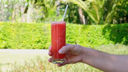 A person is sipping watermelon juice through a straw from a cylinder-shaped glass. The drink is magenta in color, made from the plant watermelon, grown in soil.