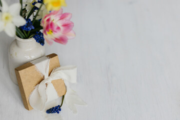 Stylish gift box with ribbon and spring flowers on rustic white table. Happy womens day and Mother's day. Beautiful daffodils and tulips gentle bouquet and present. Space for text