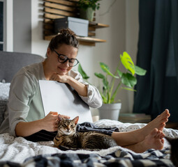 Young woman with cute gray kitten working on a project using laptop in bed at home - 750088159