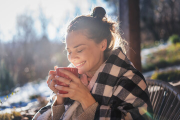 Young woman enjoying a sunny early spring morning on the terrace, wrapping herself in a blanket and drinking hot drink from red cup - 750088312