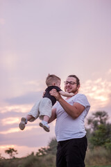 Young, diversified father throws little son into air against backdrop of soft pastel sky at sunset. Toddler is gleefully thrown in the air by parent, with background dusk, symbolizing joy and freedom