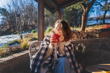 Young woman enjoying a sunny early spring morning on the terrace, wrapping herself in a blanket and drinking hot drink - 750090370