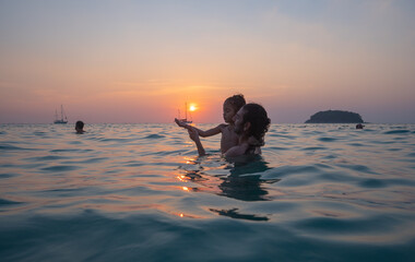 A father carries his child to play in sea water by the beach at sunset. .The father is taking the opportunity to bond with his child and enjoy a peaceful evening. .The sun is on the palm of his hand.