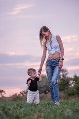 Little boy gleefully taking first steps in green field, guided by young mother, with soft sunset hues painting the sky. Woman and child walking in the countryside. Mothers day