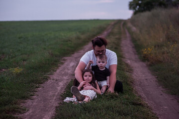 Fototapeta na wymiar In the dimming light of dusk, father sits on rural trail, his children nestled in his arms, picture of protective love, quiet contemplation. Little girl and small boy playing near man