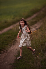 Little girl with long hair in a dress runs across a green field. Active, fun childhood. Journey to the meadow in the countryside. Ease, freedom of life. Travel weekend. Trodden path. Nature outside