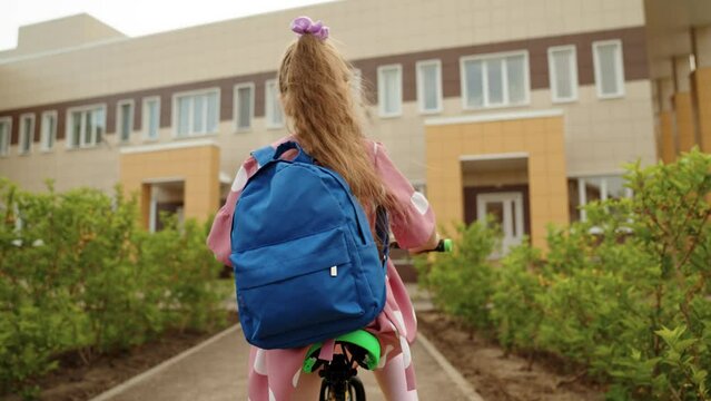 Active cute little girl pupil with backpack driving cycle arriving to elementary school campus back view closeup. Female kid child riding bike on schoolyard enjoy happy childhood sport leisure hobby