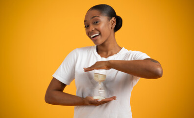 Positive Black Lady Holding And Showing Sand Clock, Yellow Backdrop
