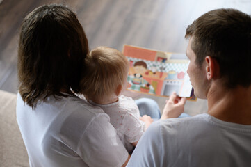 Mom and dad read an educational book to a child, sitting on the sofa, looking at pictures, rear view. Young loving parents spend time with their baby. Concept of happy family, learning. Children's Day