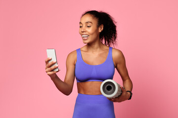 Athletic African woman holds smartphone and foam roller, pink background