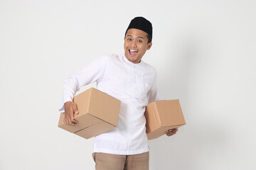 Portrait of attractive Asian muslim man in koko shirt with peci feeling grateful and peacful while carrying cardboard box. Going home for Eid Mubarak. Isolated image on white background