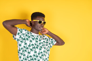 Young black man in yellow sunglasses and summer t-shirt with palm trees on a yellow background with copy space.