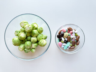 Fresh Brussels sprouts in glass bowl next to bowl of colorful candy on white background. Healthy versus indulgent food concept.
