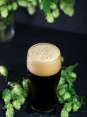 Foamy dark beer glass on the table with green hops. Craft strong stout beer
