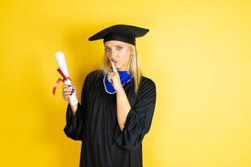Beautiful blonde young woman wearing graduation cap and ceremony robe asking to be quiet with...
