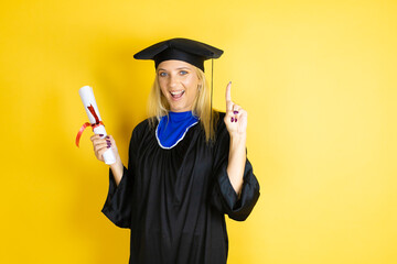 Beautiful blonde young woman wearing graduation cap and ceremony robe amazed and surprised looking at the camera and pointing up with fingers and raised arms