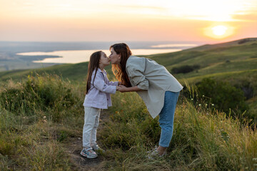 Fototapeta na wymiar Young mother kissing her baby girl daughter at sunset on the background of the lake. Family holiday on pond. Portrait mom with child together in nature. Happy Mothers Day.
