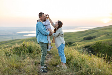 Happy family concept . Little girl is sitting on her father's shoulders, lake on the background. Young mother carries her baby girl daughter at sunset. Family holiday in nature.