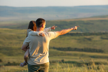 Fototapeta na wymiar Young father holds his daughter in his arms and points into the distance. Father Day. Happy man with little daughter spending time together and having fun at summer sunset.