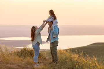 Happy family: mother father and child daughter on nature on sunset. The child sits on his father's shoulders. Summer concept. People spend time outdoor in summer