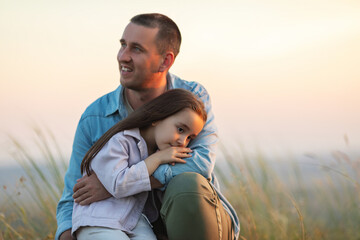 Portrait of happy young dad and his cute little daughter. Girl is sitting on her father's arms, sky and nature on the background