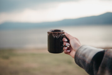 Close-up of a hand holding a dark ceramic mug with a scenic lake and mountain range in the...