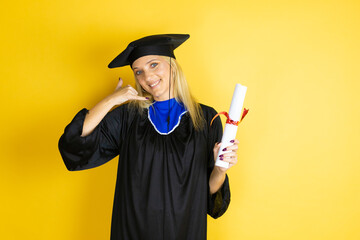 Beautiful blonde young woman wearing graduation cap and ceremony robe smiling doing phone gesture with hand and fingers like talking on the telephone