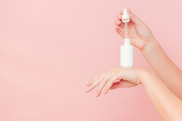Hands holding glass bottle with dropper lid. White container with cosmetic product, serum (essential oil ) on pink background. Concept of beauty