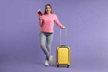 Happy young woman with passport, ticket and suitcase on purple background