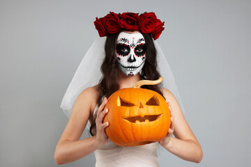 Young woman in scary bride costume with sugar skull makeup, flower crown and carved pumpkin on...