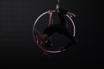 Young woman performing acrobatic element on aerial ring against dark background
