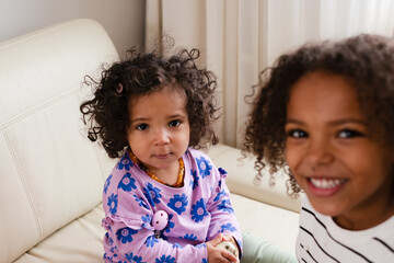 A African american young girl is smiling happily beside her sad and crying toddler sister, both...