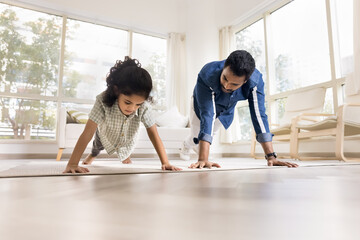Athletic strong Indian dad and little daughter kid doing pushups, exercising at home, caring for...