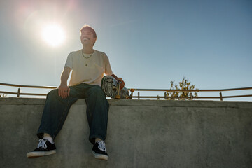 Low angle of young male skater in casual outfit sitting on ramp with skateboard against blue sky