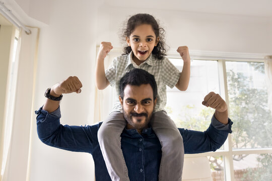 Cheerful strong Indian dad lifting little daughter kid on shoulders, making power hands gestures, showing fists, muscles, looking at camera with martial scream, playing active games