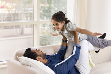 Happy young Indian dad lying on back, holding little daughter girl on arms and legs, lifting child up in air for playing airplane, laughing, having fun, enjoying leisure, fatherhood, doing acroyoga