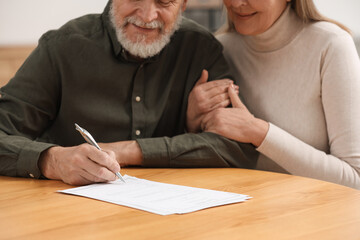 Senior couple signing Last Will and Testament at wooden table indoors, closeup