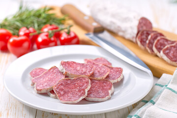fuet sausage cut in slices on a white plate with peppercorns and fresh rosemary, on a white wooden table.