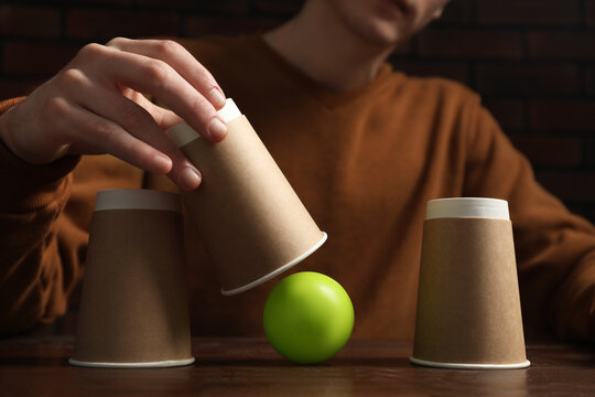 Shell game. Man showing ball under cup at wooden table, closeup