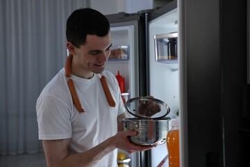 Man with sausages and pot near refrigerator in kitchen at night