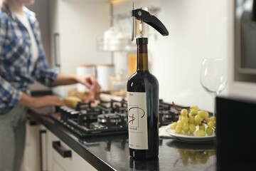 Wine bottle with corkscrew on black countertop, closeup. Woman in kitchen, selective focus