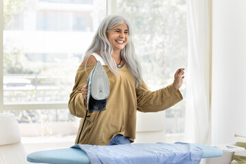 Positive joyful senior Latin woman having fun while doing housework at home, holding hot iron over board with dry cloth, dancing to funny music, laughing, smiling, looking away