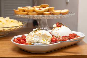 A plate of cream cheese with tomatoes and olive oil, with a platter of gourmet empanadas and assorted cheeses in the background
