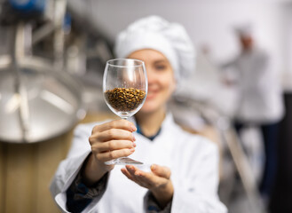Closeup of glass with malted barley grain in hands of smiling female brewmaster, selective focus....