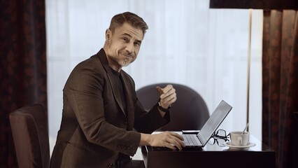 Mature businessman working late on laptop computer at home, coffee cup on table. Portrait of happy man sitting at desk in hotel room or at home managing tasks online.