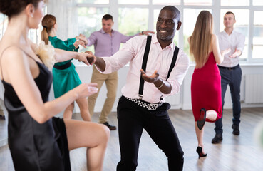 Woman in elegant evening dress dances incendiary boogie woogie with African American man partner during choreography lesson. Group training and rehearsal, preparation for competitions