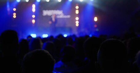 The view from the auditorium, on a concert stage with a professional stage light in blue tones, a speaker on personal growth or success speaks. Unrecognizable faces. High quality 4k footage