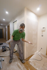 Worker wearing face mask sanding plasterboard walls covered with dust all over his body. Dirty work